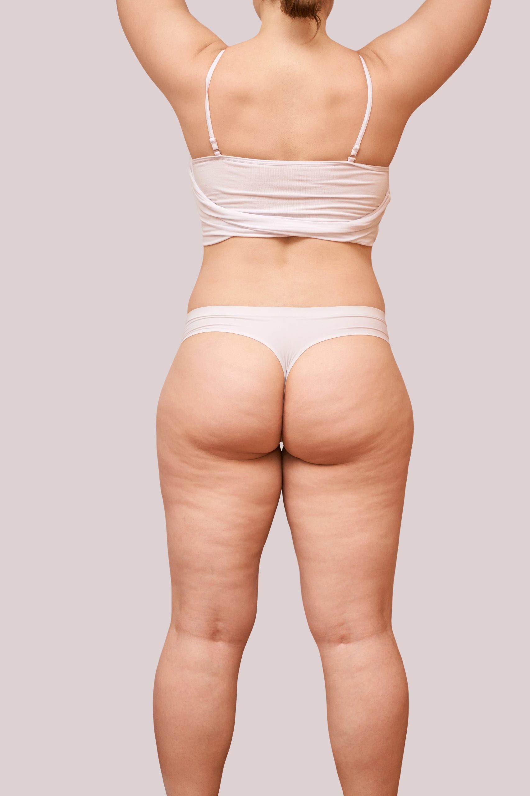 Caucasian female with undergarment displays natural body from rear view on neutral background.